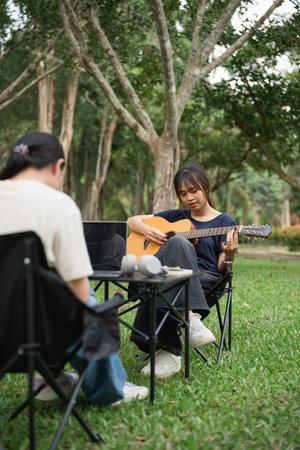 Young woman playing acoustic guitar in the park while friend sits at table with laptop and speakers, enjoying leisurely outdoor music sessionの写真素材
