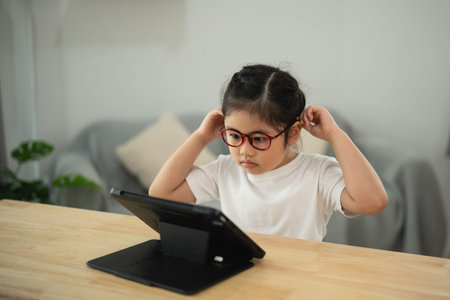 Young girl wearing stylish eyeglasses focused on tablet screen while sitting at wooden table in...