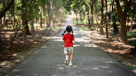 Young Girl in Red Shirt Running on a Tree-Lined Path in a Scenic Park Surrounded by Green Trees and Nature, Focus on Childhood Joy and Activityの写真素材