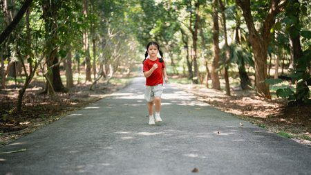 Young girl running joyfully on a path surrounded by green trees in a serene park setting, enjoying the beauty of nature and playfulness of childhoodの写真素材