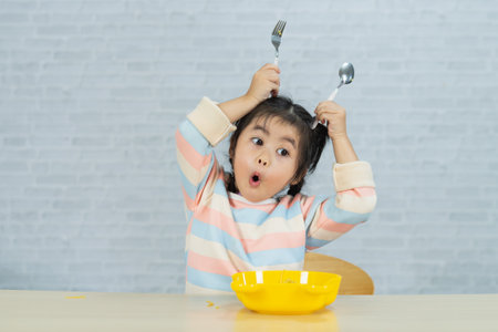 Excited young girl playing with utensils in a joyful and creative expression, showcasing innocence and fun during mealtime activities at homeの写真素材