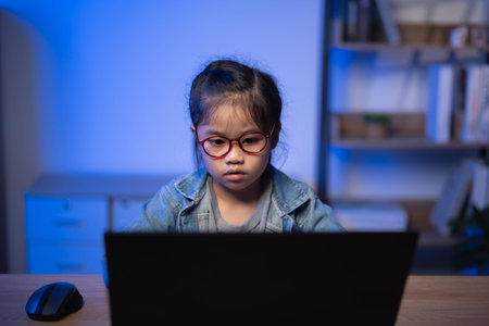Young girl focused on computer screen in cozy room with blue lighting, wearing glasses and denim...