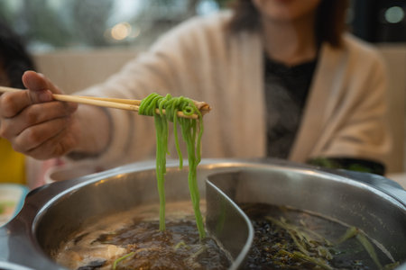 A woman expertly holds noodles with chopsticks above a bubbling hot pot, showcasing a delicious meal...