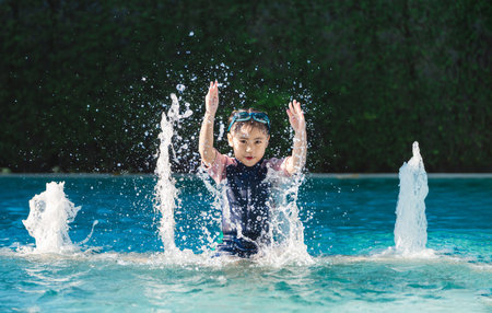 Happy child playing in swimming pool, splashing water with hands raised, enjoying summer day under bright sunlight with playful energy and joyの写真素材
