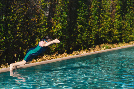 Child in Swim Gear Leaps into Clear Pool Water with Lush Greenery in Background Capturing Joy of Summer Fun and Outdoor Activitiesの写真素材
