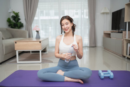 Young woman stretches in a modern living room while sitting on a yoga mat with dumbbells and a water bottle, promoting health and wellness lifestyleの写真素材