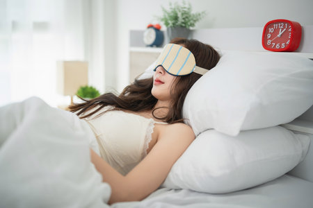 Woman Relaxing in Bed with Eye Mask, Peaceful Sleep Environment, Cozy Bedroom with Bright Light and Soft Bedding Accentsの写真素材