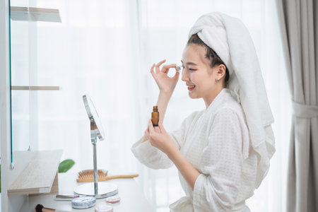 Woman with towel on head applying skincare serum in bright bathroom setting, enjoying self-care routine and beauty therapy at homeの写真素材