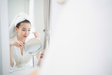 Young woman in white bathrobe with towel on head applying skincare products in front of mirror in bright, modern bathroom settingの写真素材