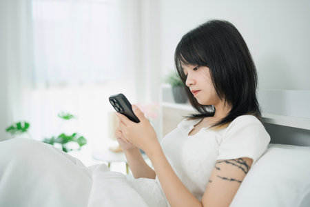 Young woman sitting in bed while using smartphone, wearing casual white clothing, surrounded by a bright and cozy bedroom environment with plantsの写真素材
