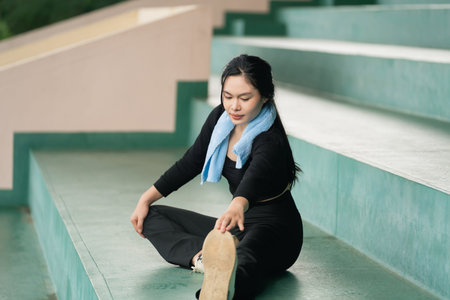 Young woman stretching on outdoor stairs wearing a black outfit and blue towel, practicing yoga in a...