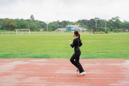 Young woman jogging on an athletic track during a cloudy day, showcasing determination and fitness in a serene outdoor environment for sports enthusiastsの写真素材