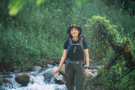 Adventurous Woman Smiling by a Flowing Stream in Lush Green Nature, Wearing Hiking Gear and Holding a Water Bottle, Embracing Outdoor Explorationの写真素材