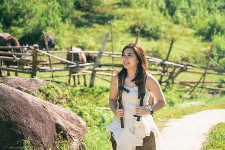 Young woman trekking through a scenic landscape with a forest background, enjoying nature, wearing backpack and casual outfit, smiling and relaxedの写真素材