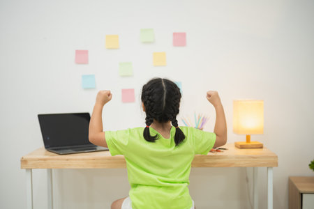 Empowering young girl with braids celebrating achievement in learning environment, colorful sticky notes on wall, laptop, and desk setup illuminated by warm lightの写真素材