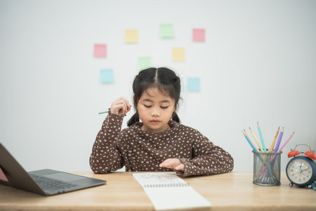 Young girl engaged in creative learning with notebook and colored pencils at a study table surrounded by sticky notes and a laptop, focused on her tasksの写真素材