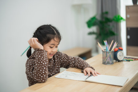 Happy young girl enjoying drawing in notebook at wooden table in cozy room with green plant and colorful pencils on a sunny day.の写真素材