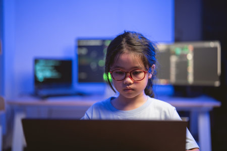 Young Girl Concentrated on Laptop Surrounded by Modern Technology in a Room with Computer Screens Displaying Code and Data Visualizationsの写真素材