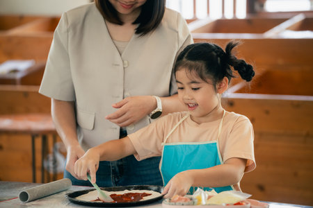 Mother and daughter enjoy cooking together in kitchen, child learns to spread sauce on pizza base, family bonding, culinary fun, and childhood momentsの写真素材