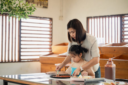 Caring mother guides young daughter in making pizza together in bright kitchen with wood accents and natural light, showcasing bonding experienceの写真素材