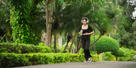 A young woman jogs through a vibrant park filled with lush greenery, wearing headphones and enjoying...