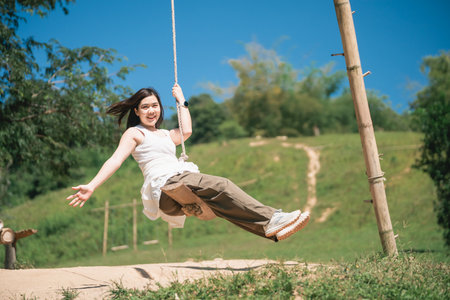 Young woman enjoying a playful swing ride on a sunny day surrounded by greenery with a bright blue sky in the background reflecting happiness and freedomの写真素材