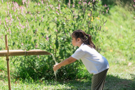 Child Playing with Water from Bamboo Fountain in a Lush Green Garden Surrounded by Flowers and Natural Beauty on a Sunny Dayの写真素材