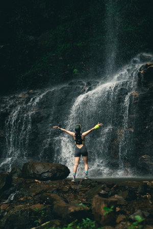 Woman Standing in Front of Majestic Waterfall with Arms Raised Celebrating Nature and Adventure in a Lush Outdoor Environmentの写真素材