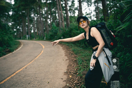 Young woman hiking in forest, wearing sporty outfit, gesturing for ride, outdoor adventure, lush scenery, nature exploration, summer travel, minimalismの写真素材