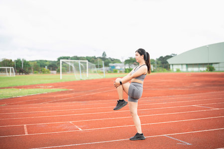 Young woman stretching on outdoor athletic track during workout session in gray sportswear,...