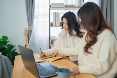 Two Women Collaborating on Creative Ideas in a Cozy Workspace with Laptop, Notebook, and Indoor Plants in Bright Natural Lightの写真素材