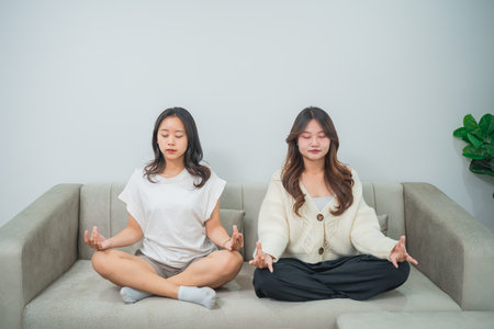 Two Women Meditating Together on Couch in Minimalistic Living Room Setting for Relaxation and Mindfulness Practicesの写真素材