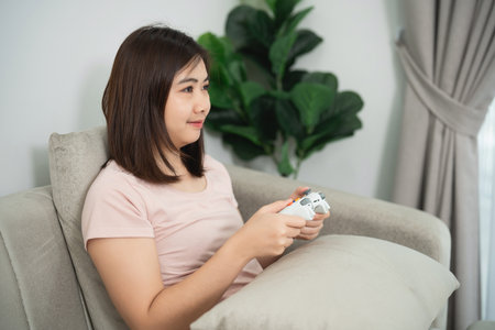 Young woman enjoying video game experience at home, cozy living room setting, relaxing on couch with controller and soft pillow in modern interiorの写真素材