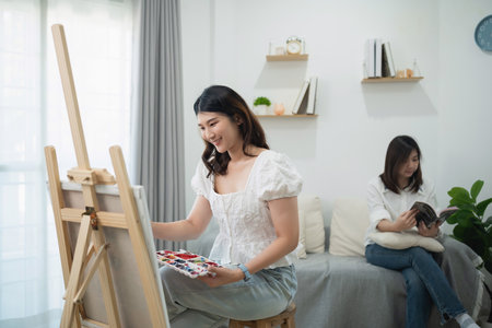 Young Woman Painting on Canvas in Cozy Living Room with Friend Reading Book on Sofa, Bright Natural Light and Minimalist Decor Settingの写真素材
