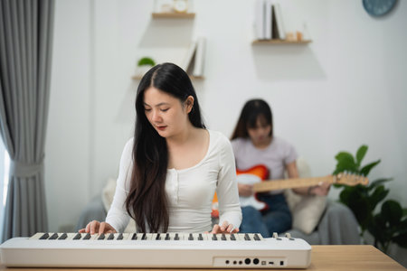 Young women play keyboard and electric guitar together in cozy living room creating music and fostering friendship in modern home environmentの写真素材