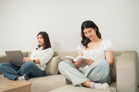 Cozy indoor scene with two young women relaxing on a couch, one reading a book and the other working...