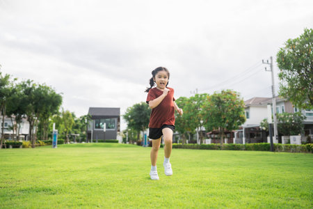 Happy young girl running energetically on green grass in sunny park, enjoying outdoor activity, showcasing vitality and childhood joy in a neighborhood settingの写真素材