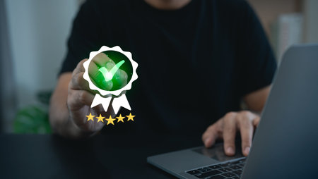 Person Holding a Green Check Mark Badge Symbolizing Achievement with Laptop on Table in Modern Office Settingの写真素材
