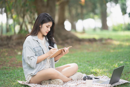 Young woman writing in notebook while sitting on grass in park with laptop and coffee cup, enjoying peaceful outdoor workspace in natureの写真素材
