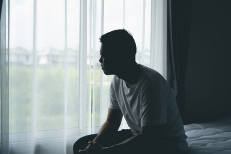 Solitary Reflection of a Young Man Contemplating Life While Seated by a Bright Window Surrounded by Soft Drapes in a Modern Interior Spaceの写真素材