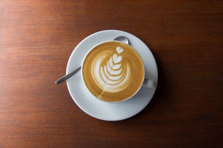 Top-view shot of single cup of latte coffee with art cream foam in white ceramic cup on wooden vintage table with sun lighting and shade in the morning after breakfast for relaxing and starting a day.の写真素材