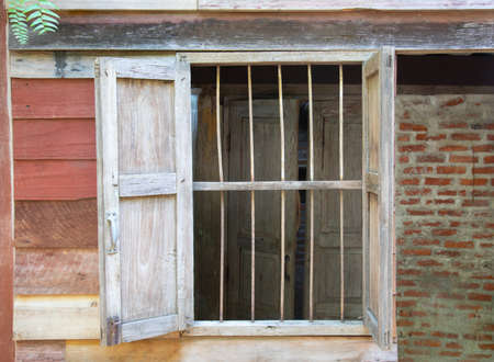 Close up shot of grunge wooden window frame in vintage and retro styles of the rural house which is opening shows beautiful texture of reddish wooden and brick walls with fern tree on the roof.の写真素材