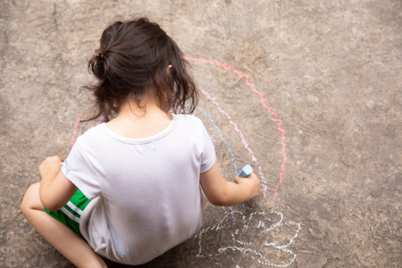 Top view shot of preschool child girl whos is drawing colorful rainbow on pavement or concrete floor in the park by the color chalk in summer time shows the expression of her imagination by activity.の写真素材