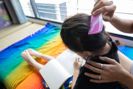 Back view of asian mom making braids for her black hair adorable child daughter in room at home shows concept of parental love and togetherness. It is building connection and family relation.の写真素材