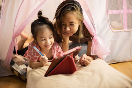 Face focusing on asian mom and adorable daughter are lying on the floor in pink tent at home and writing on tablet which is using digital technology for happy and joyful moment together.の写真素材