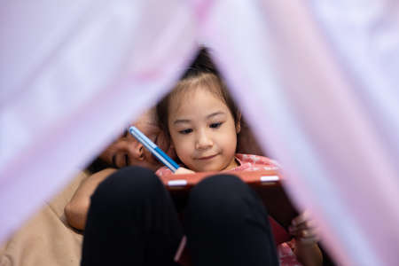 Sneaking on asian mom and adorable daughter are laying on the floor in the house and paying happy and joyful moment together by talking and writing on tablet which is use technology for fun.の写真素材