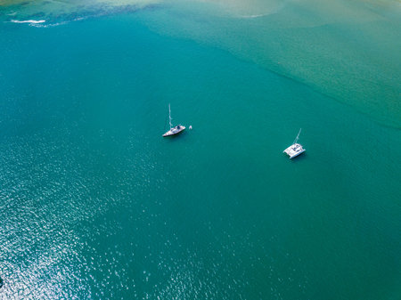 A view from a high-angle drone sees sailboats leaving anchors in a beautiful bay, clear glass-like...