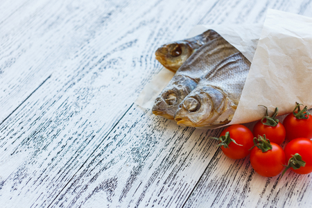 Three dried fish bream lie on a light wooden table. Fish wrapped in a paper bag. Near to lay five cherry tomatoes.の写真素材