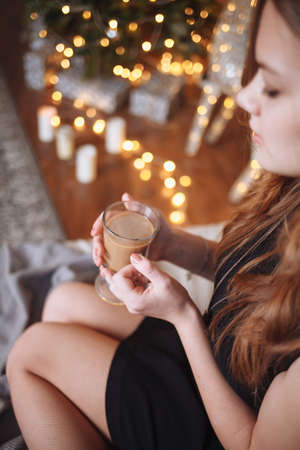 Portrait of an attractive young girl in a black evening dress on the bed against the background of a dressed up Christmas tree with a cup of cocoa in her hands.の写真素材