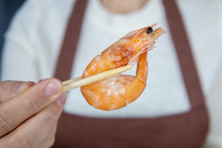 A girl in a white blouse and brown apron holds a large red king prawn with chopsticks. Asian food concept.の写真素材